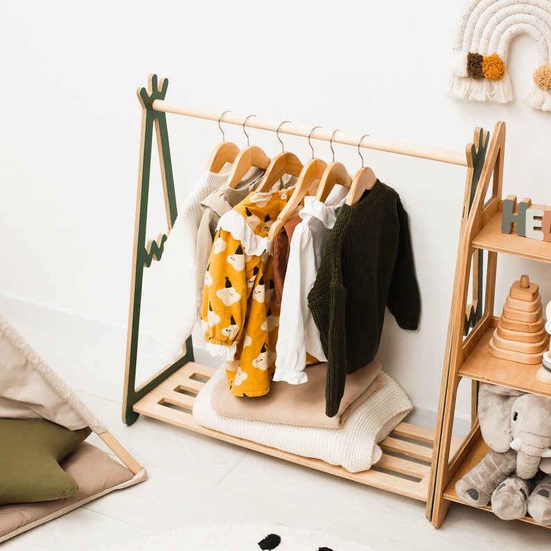 Puede incluir: Un perchero de madera con un dise&ntilde;o verde y marr&oacute;n, que incluye una barra para colgar y un estante. El perchero est&aacute; decorado con una variedad de ropa para ni&ntilde;os, incluyendo una camisa con estampado amarillo y negro, una camisa verde y una camisa blanca.