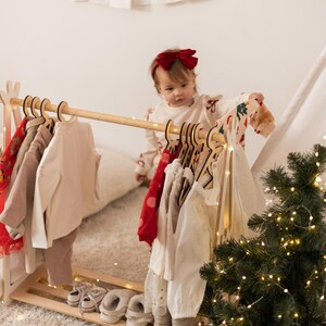 May include: A wooden clothes rack with a variety of clothing items hanging on it. The rack is decorated with a string of lights. A young child is standing in front of the rack, holding a red and white striped shirt. The child is wearing a red bow in their hair.