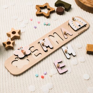 以下が含まれることがあります： パステルカラーの文字で「EMMA」と書かれた木製の名前パズルボード。蝶の形をしたピースと虹のアーチのピースが含まれています。各文字と形には、掴むための小さな木製のノブが付いています。ボードにはChildUniverseのブランド名が記されています。