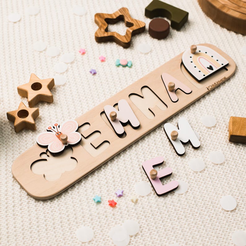 以下が含まれることがあります： パステルカラーの文字で「EMMA」と書かれた木製の名前パズルボード。蝶の形をしたピースと虹のアーチのピースが含まれています。各文字と形には、掴むための小さな木製のノブが付いています。ボードにはChildUniverseのブランド名が記されています。
