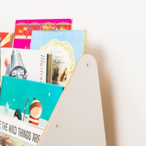 May include: A white wooden bookshelf with a triangular shape, holding a variety of colorful children's books. The books have titles like "The Complete Adventures of Rabbit" and "Where the Wild Things Are".