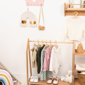 May include: A wooden clothing rack with a pink and white rainbow macrame hanging above it. The rack is filled with children's clothing, including a white dress, a pink and white gingham dress, a green and white polka dot dress, and a green sweater. There are also two pairs of white shoes and a pair of brown shoes on the rack. The rack is in a white room with a wooden shelf with a rainbow abacus on it.