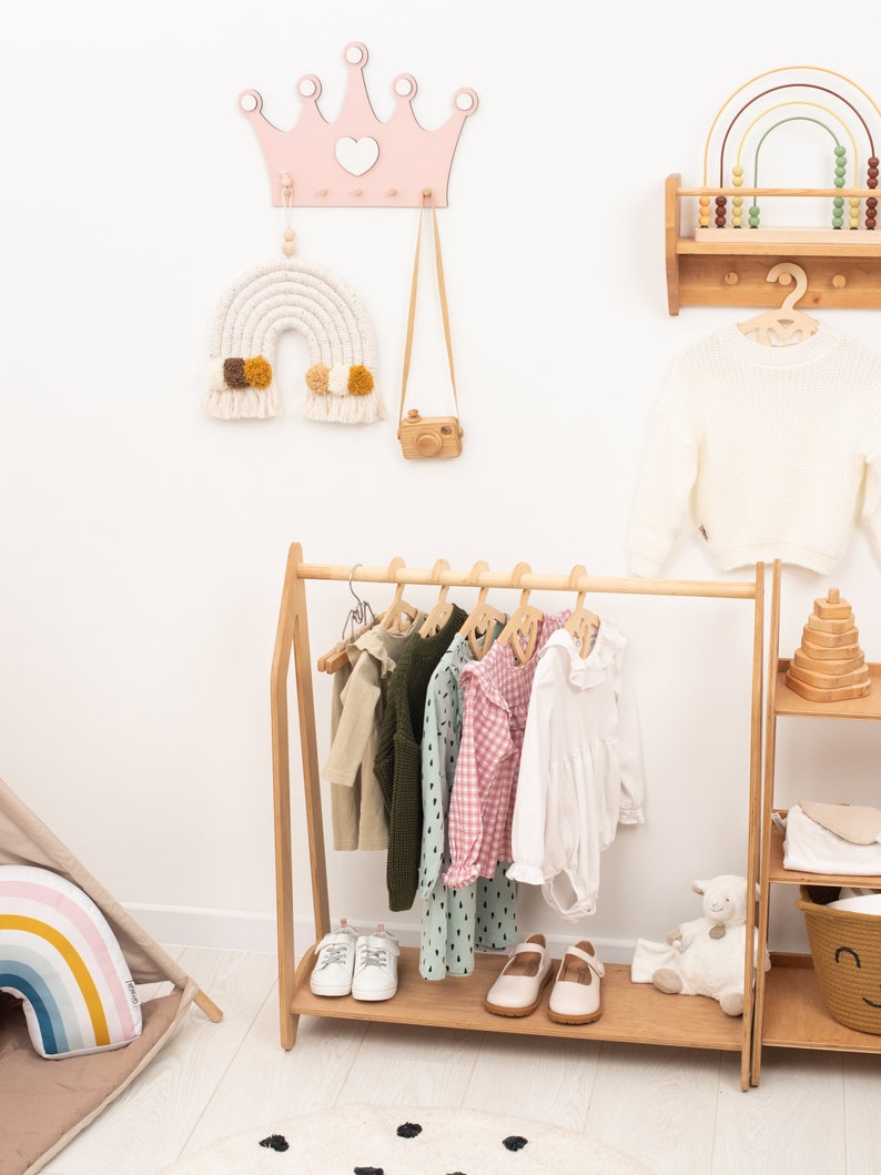 May include: A wooden clothing rack with a pink and white rainbow macrame hanging above it. The rack is filled with children's clothing, including a white dress, a pink and white gingham dress, a green and white polka dot dress, and a green sweater. There are also two pairs of white shoes and a pair of brown shoes on the rack. The rack is in a white room with a wooden shelf with a rainbow abacus on it.