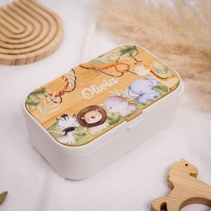 May include: A white rectangular lunchbox with a bamboo-style lid featuring jungle animals. The lid has a zebra, lion, elephant, and giraffe, with the name "Olivia" in cursive. The lunchbox is decorated with green leaves and vines.