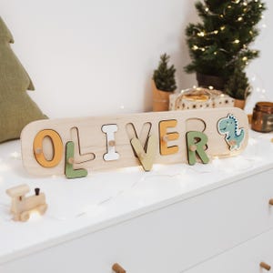 以下が含まれることがあります： 切り抜かれた文字で「OLIVER」と書かれた木製の名前パズルボード。文字はマスタードイエロー、セージグリーン、ホワイトで彩色されています。明るい木製のボードの右側には、小さな漫画の恐竜のフィギュアが取り付けられています。