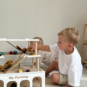 K&ouml;nnte beinhalten: Ein mehrst&ouml;ckiges Parkhaus aus Holz mit Rampen. Das Parkhaus ist wei&szlig; und tr&auml;gt den Schriftzug "My Parking". Verschiedene Spielzeugautos und Baufahrzeuge sind auf den Ebenen angeordnet. Zwei Kinder spielen mit dem Spielzeug.