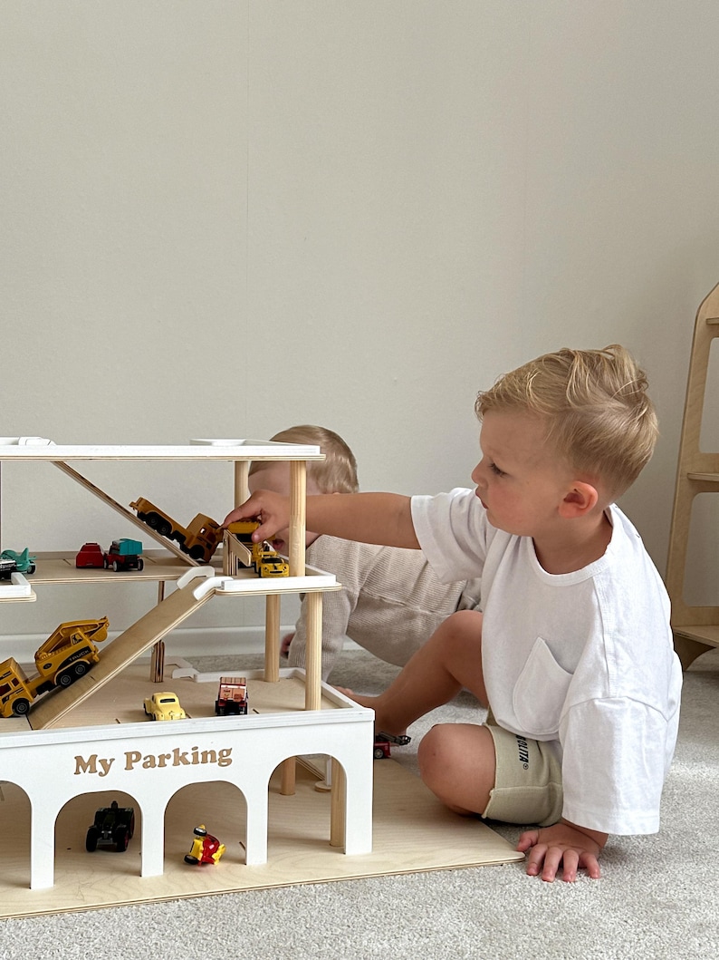 K&ouml;nnte beinhalten: Ein mehrst&ouml;ckiges Parkhaus aus Holz mit Rampen. Das Parkhaus ist wei&szlig; und tr&auml;gt den Schriftzug "My Parking". Verschiedene Spielzeugautos und Baufahrzeuge sind auf den Ebenen angeordnet. Zwei Kinder spielen mit dem Spielzeug.