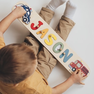 May include: Wooden name puzzle with the name "JASON" in colorful, raised letters. The puzzle features a police car and a fire truck graphic on each end. The child is wearing a yellow shirt and khaki pants.