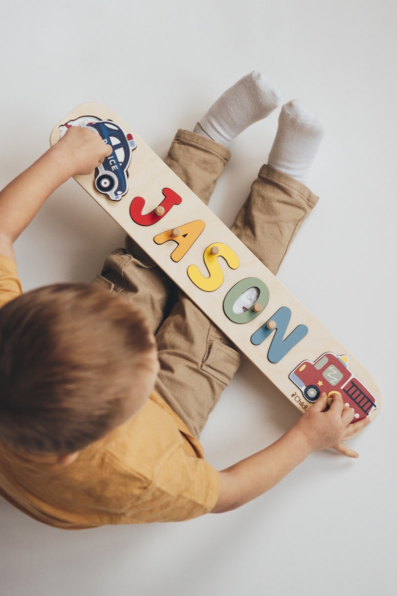 May include: Wooden name puzzle with the name "JASON" in colorful, raised letters. The puzzle features a police car and a fire truck graphic on each end. The child is wearing a yellow shirt and khaki pants.
