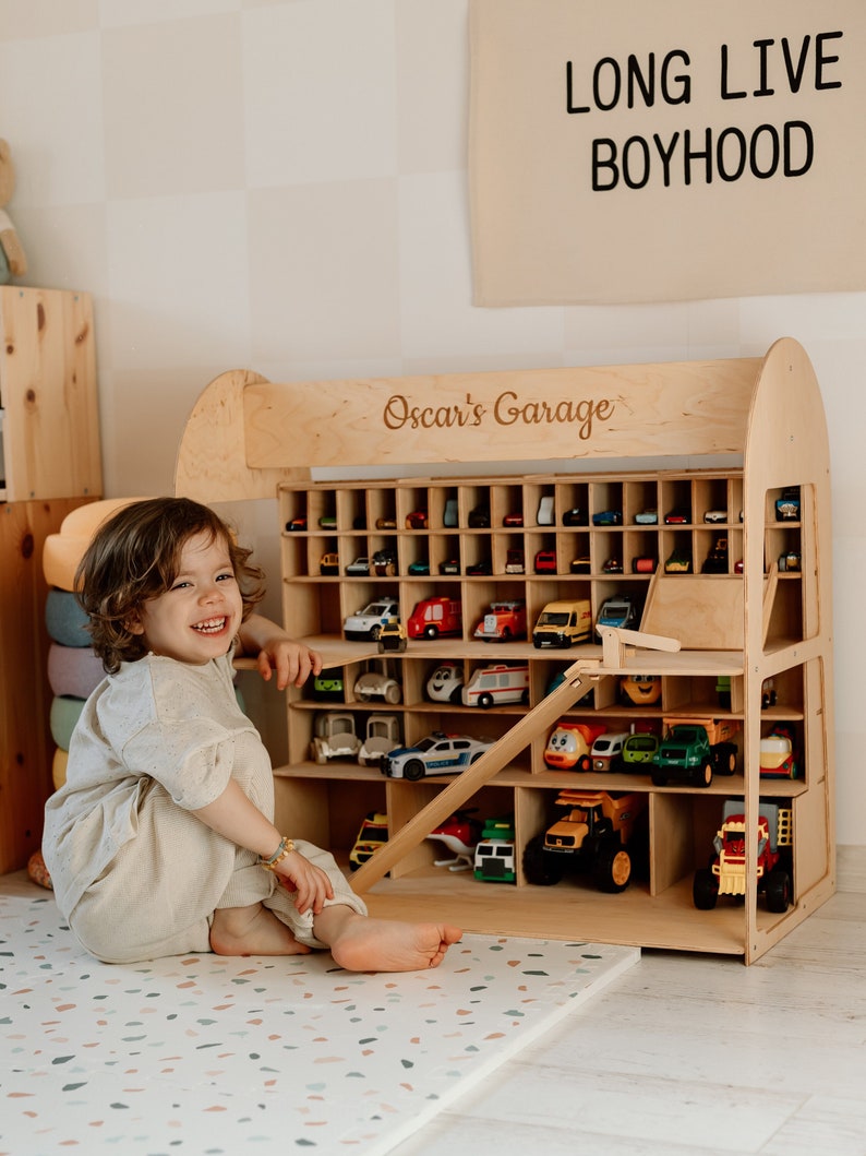 K&ouml;nnte beinhalten: Ein Holz-Spielzeug-Autogarage mit vielen kleinen Spielzeugautos. Auf der Garage steht "Oscar's Garage". Ein Banner dar&uuml;ber zeigt "LONG LIVE BOYHOOD".