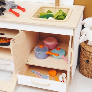 May include: A white wooden play kitchen with an open cabinet and drawer. The cabinet contains a blue, pink, and light blue toy pots and pans. The drawer contains a variety of toy kitchen utensils.