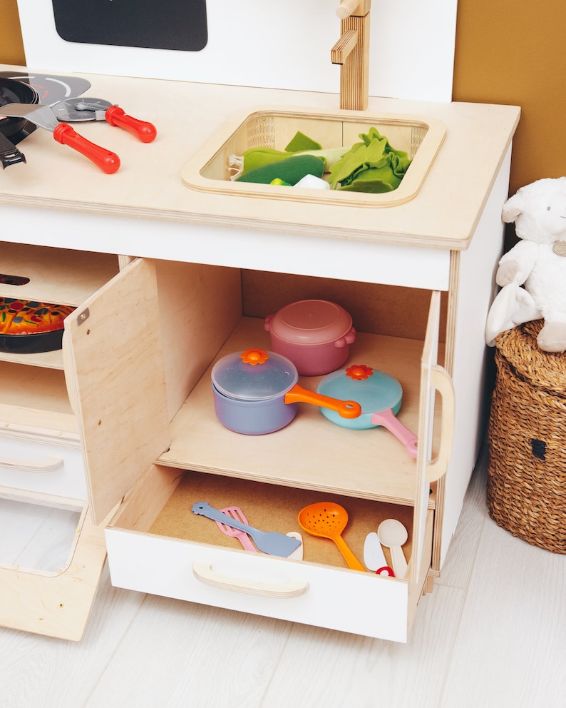 May include: A white wooden play kitchen with an open cabinet and drawer. The cabinet contains a blue, pink, and light blue toy pots and pans. The drawer contains a variety of toy kitchen utensils.