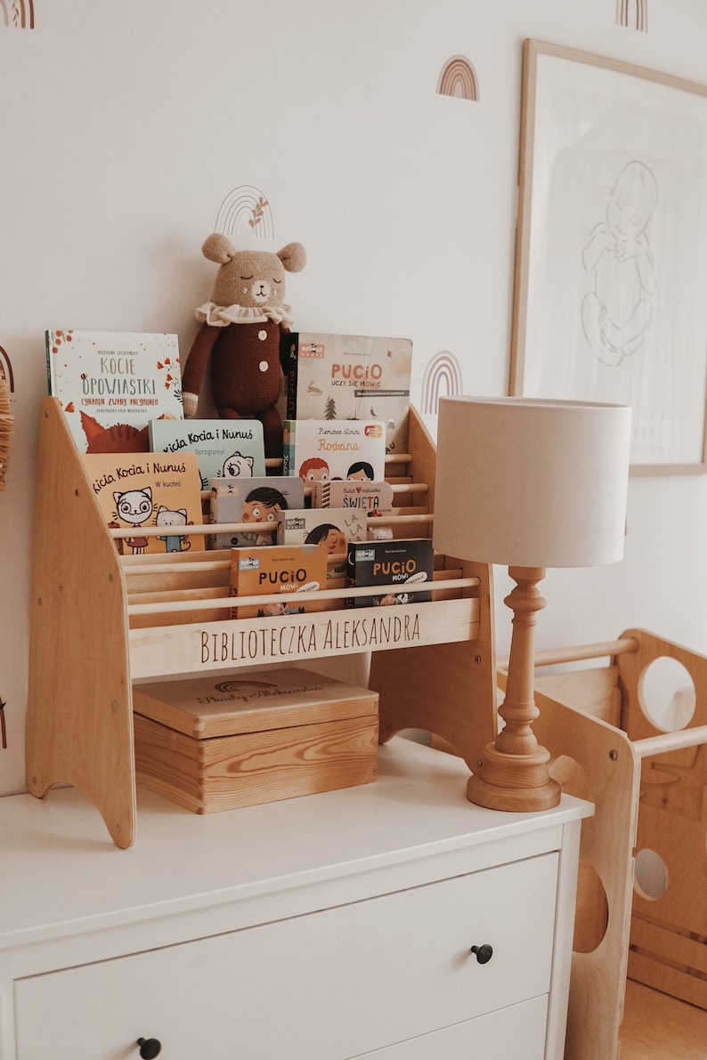 May include: A wooden bookshelf with a light wood finish, labeled "Biblioteczka Aleksandra" and filled with children's books. A stuffed bear sits on top of the bookshelf. A white lamp with a beige shade sits on a white dresser next to the bookshelf.