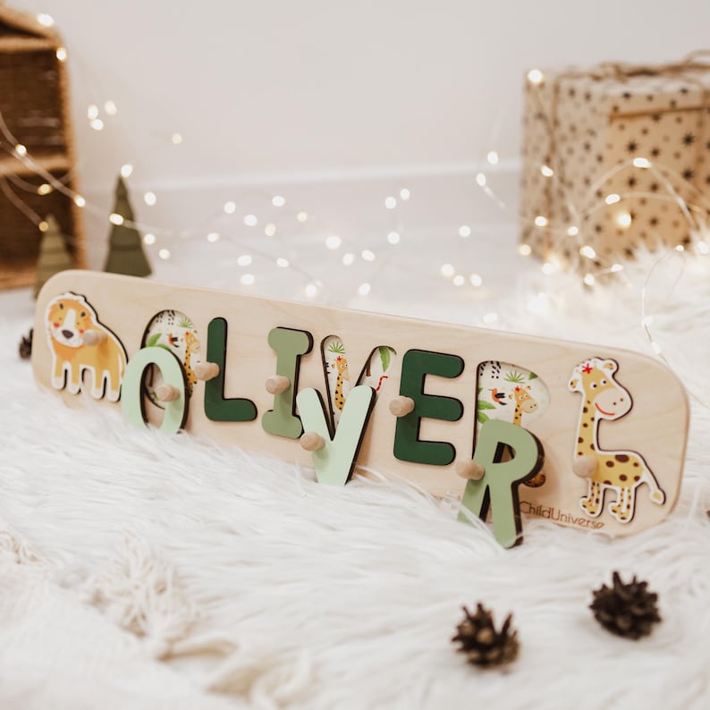 May include: Wooden puzzle featuring the name "OLIVER" with animal illustrations. The letters are in green and light green, with animal-themed cutouts. A dog and giraffe are on either end. The puzzle is made of light-colored wood.