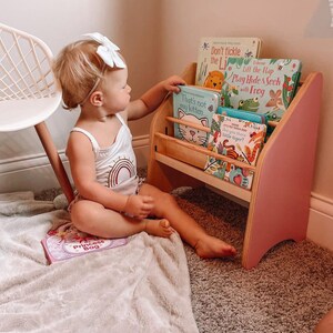 May include: A wooden children's bookshelf with pink side panels and horizontal shelves holding several board books, including titles like Don't tickle the Lion, Play Hide & Seek with Frog, That's not my kitten, and Are you there little tiger?