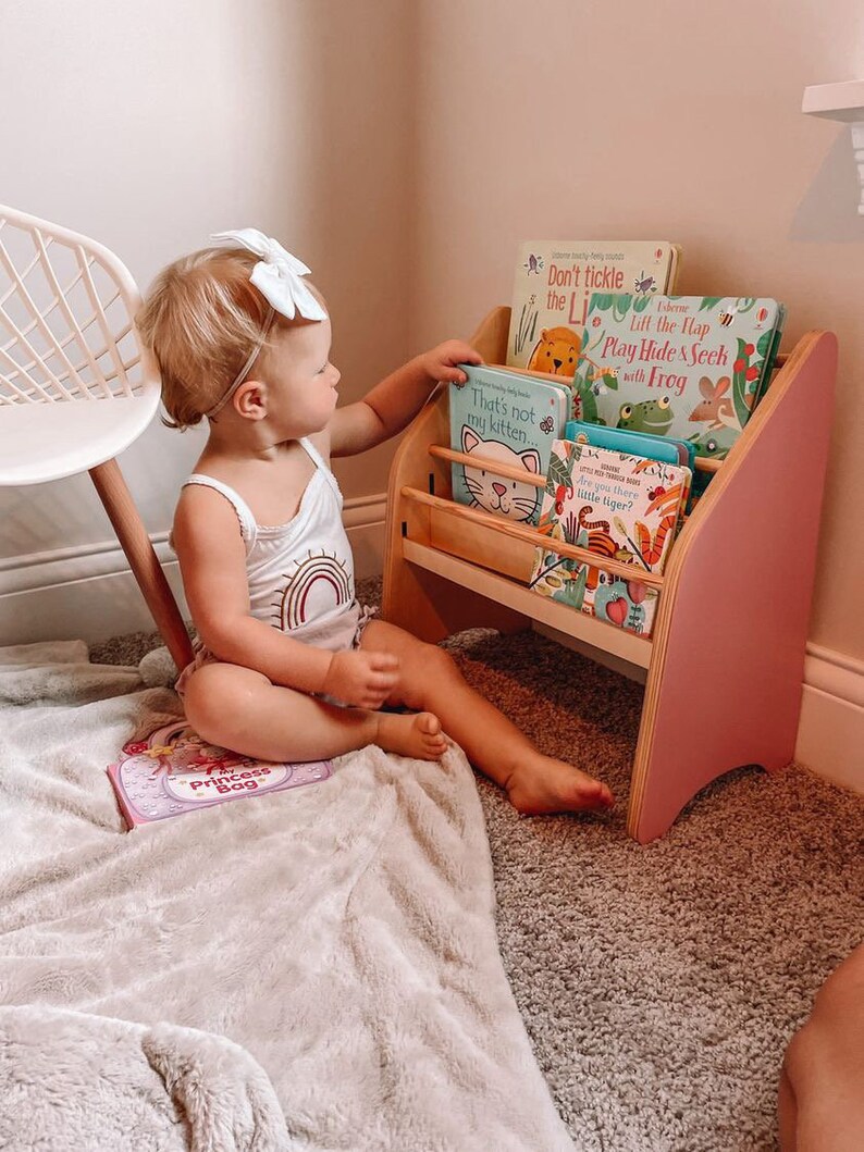 May include: A wooden children's bookshelf with pink side panels and horizontal shelves holding several board books, including titles like Don't tickle the Lion, Play Hide & Seek with Frog, That's not my kitten, and Are you there little tiger?
