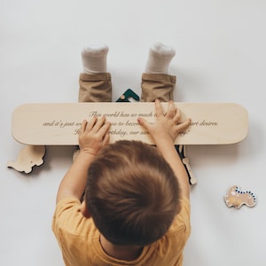 May include: A child holds a light wooden skateboard-shaped plaque with engraved text. The plaque is surrounded by wooden dinosaur cutouts and a green geometric shape. The child wears a yellow shirt and brown pants.