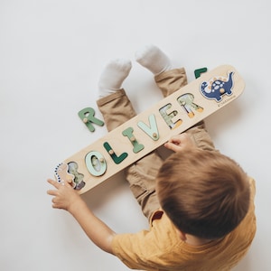 May include: A wooden name puzzle with the name "OLIVER" in green letters. The puzzle features dinosaur illustrations and is held by a child wearing a yellow shirt and khaki pants. The puzzle is approximately 12 inches long.