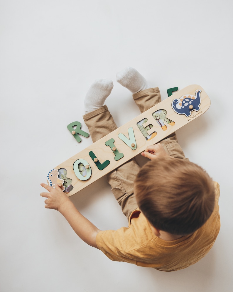 May include: A wooden name puzzle with the name "OLIVER" in green letters. The puzzle features dinosaur illustrations and is held by a child wearing a yellow shirt and khaki pants. The puzzle is approximately 12 inches long.