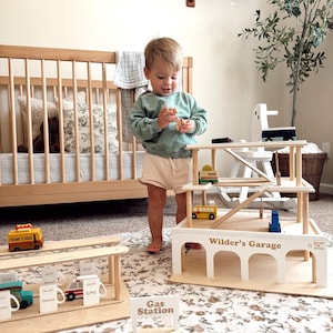 K&ouml;nnte beinhalten: Ein Holzspielzeug-Garagenset mit Tankstelle und mehrst&ouml;ckigem Parkhaus. Das Set enth&auml;lt Miniaturfahrzeuge und Schilder. Die Garage ist mit "Wilder's Garage" beschriftet. Ein Kind spielt mit einem Holzspielzeug.