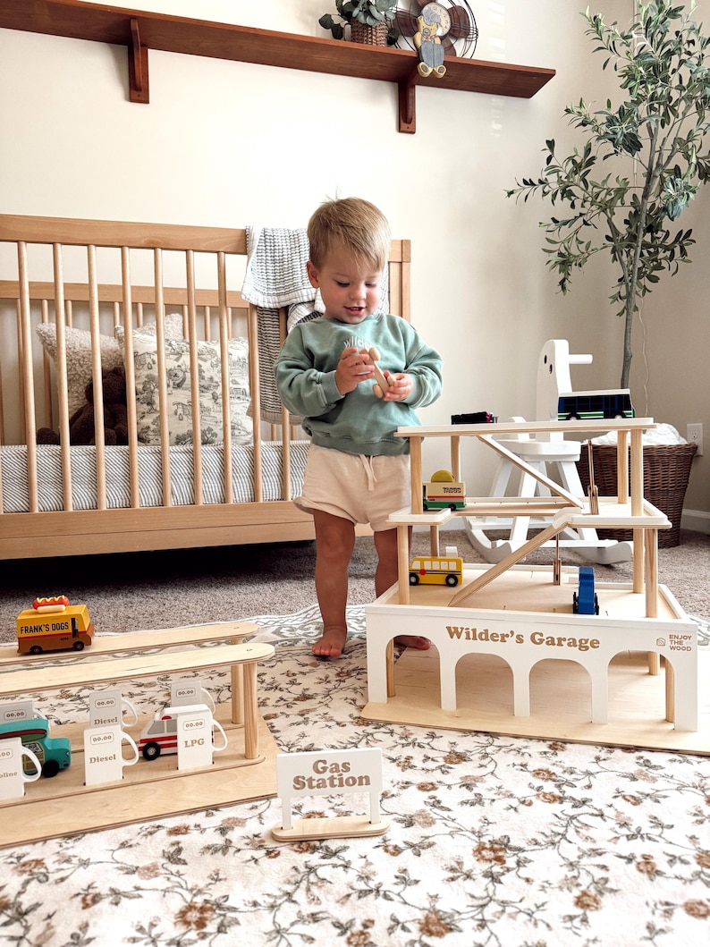 K&ouml;nnte beinhalten: Ein Holzspielzeug-Garagenset mit Tankstelle und mehrst&ouml;ckigem Parkhaus. Das Set enth&auml;lt Miniaturfahrzeuge und Schilder. Die Garage ist mit "Wilder's Garage" beschriftet. Ein Kind spielt mit einem Holzspielzeug.