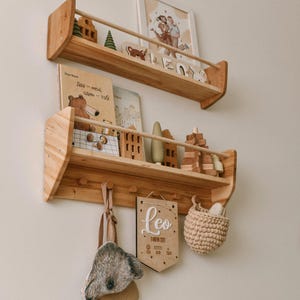 May include: Wooden wall shelves with a light finish, displaying children's books, wooden toys, and decorative items. A framed photo and a banner with the name "Leo" are also visible. A koala bear-shaped bag and a small woven basket hang below.
