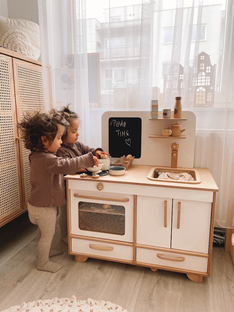 May include: Two children play with a white wooden toy kitchen playset. The kitchen has a stove, oven, sink, and cabinets. The children are wearing brown shirts and pants. The kitchen has a chalkboard sign that says "Pasta Time".