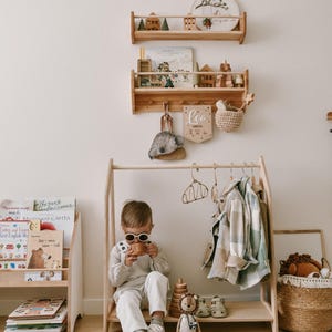 May include: A wooden children's clothing rack with a child wearing sunglasses and a white outfit sitting on the bench. The rack has a wooden shelf with a stuffed lion, a wooden ring stack, and a pair of sandals. There are also wooden shelves on the wall with books and toys.