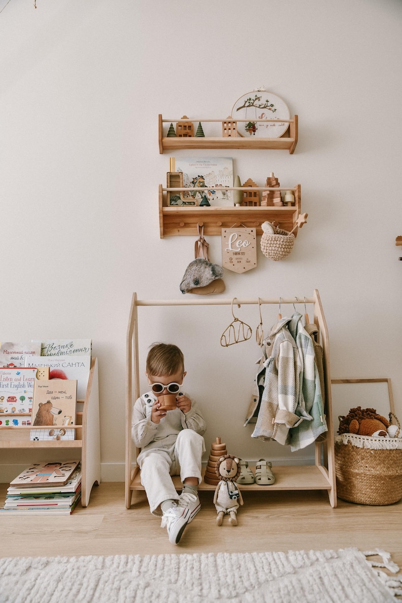 May include: A wooden children's clothing rack with a child wearing sunglasses and a white outfit sitting on the bench. The rack has a wooden shelf with a stuffed lion, a wooden ring stack, and a pair of sandals. There are also wooden shelves on the wall with books and toys.