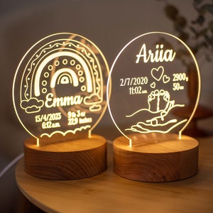 May include: Two personalised LED night lights with wooden bases. One features a rainbow design and the name "Emma", with birth details: 15/4/2023, 6:12 a.m., 9 lbs 3 oz, and 58.17 cm. The other has the name "Ariia" and birth details: 2/7/2020, 11:02 a.m., 2900g, and 50cm.