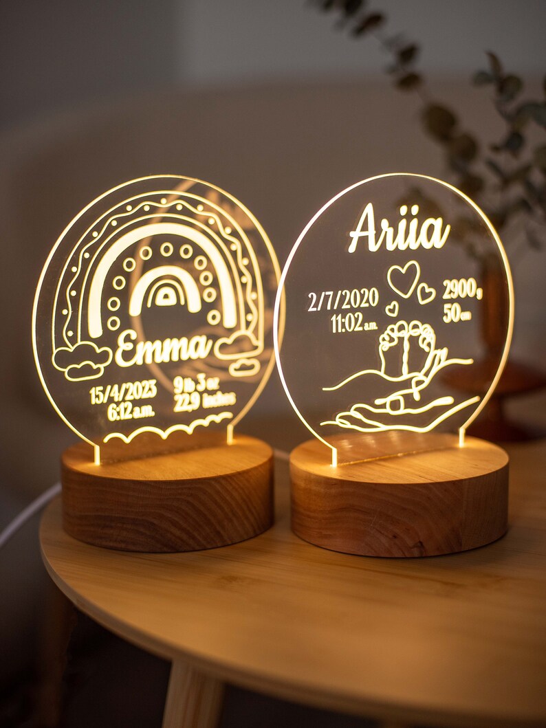 May include: Two personalised LED night lights with wooden bases. One features a rainbow design and the name "Emma", with birth details: 15/4/2023, 6:12 a.m., 9 lbs 3 oz, and 58.17 cm. The other has the name "Ariia" and birth details: 2/7/2020, 11:02 a.m., 2900g, and 50cm.