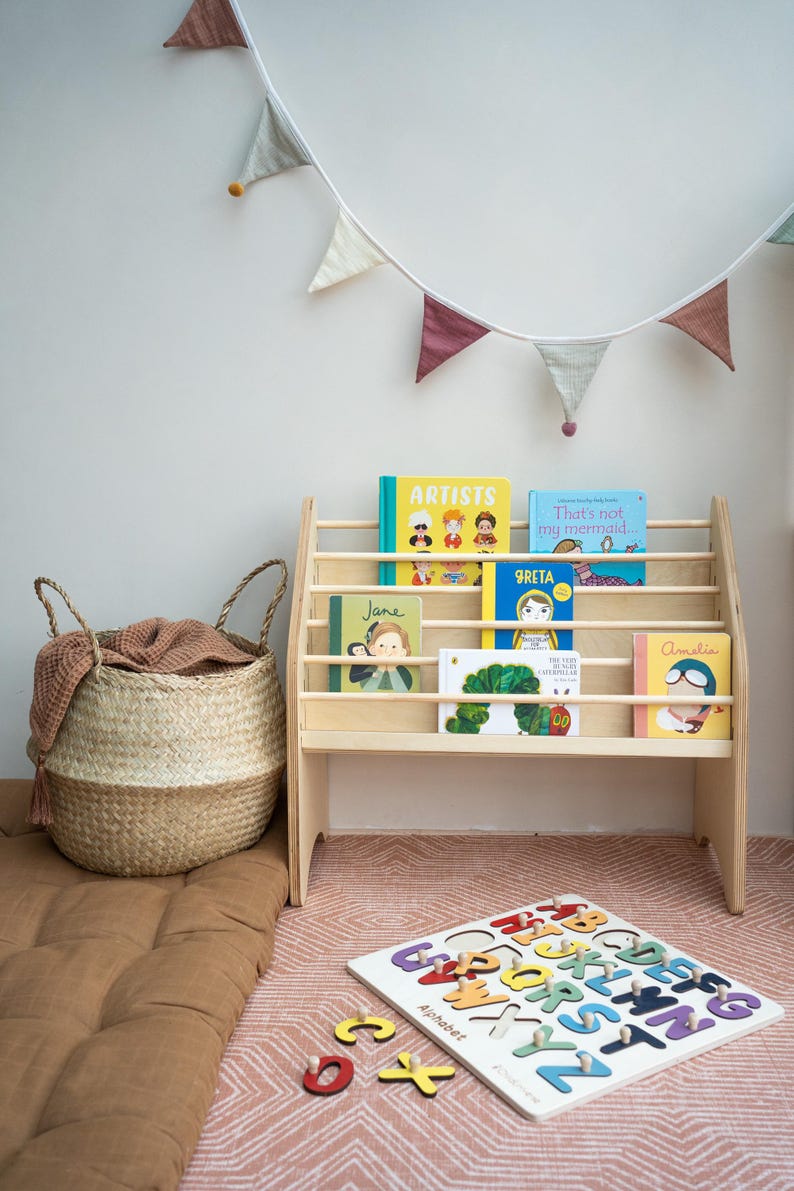May include: A light-colored wooden bookshelf holding children's books with colorful covers, a woven seagrass basket with a brown cloth, and a wooden alphabet puzzle board with individual letter pieces scattered nearby.