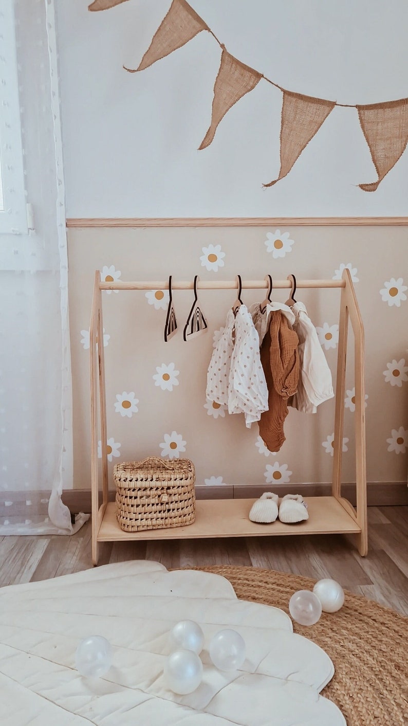May include: A wooden clothes rack with a shelf and a wicker basket. The rack is decorated with white daisy decals and a burlap banner. There are white ball pit balls on a white rug in the foreground.