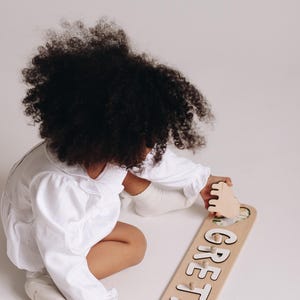 May include: A wooden name puzzle with the name "GRETA" in white letters. The puzzle has a light brown background with a cartoon leopard illustration. The child is wearing a white outfit.