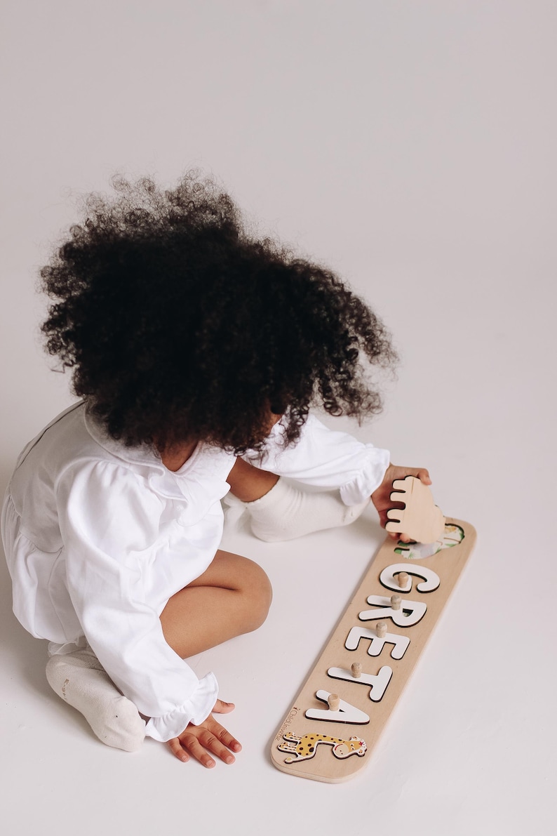 May include: A wooden name puzzle with the name "GRETA" in white letters. The puzzle has a light brown background with a cartoon leopard illustration. The child is wearing a white outfit.