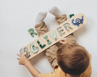 May include: A child sits on the floor playing with a wooden name puzzle. The puzzle is shaped like a long rectangle and has the name "Oliver" spelled out in green letters. The child is holding a dinosaur-shaped piece in their hand. The puzzle is made by Childhome.
