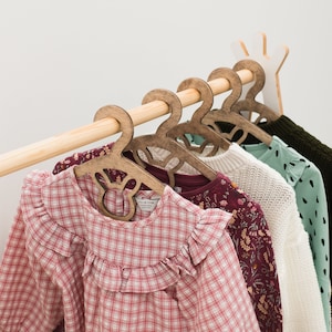 Puede incluir: Un perchero de madera con cinco perchas de madera que sostienen varias camisas estampadas. Las camisas son en tonos de rosa, rojo, blanco y verde.