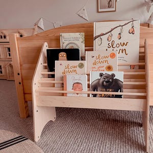 May include: A light-colored wooden bookshelf holding several children's books, including titles like Slow Down, Gandhi, and The Very Cranky Bear. The books feature illustrations of butterflies, an owl, and a bear, displayed on horizontal wooden slats.