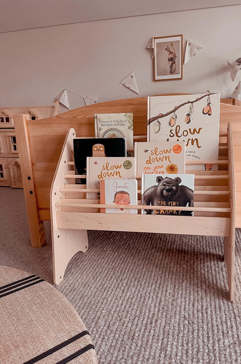 May include: A light-colored wooden bookshelf holding several children's books, including titles like Slow Down, Gandhi, and The Very Cranky Bear. The books feature illustrations of butterflies, an owl, and a bear, displayed on horizontal wooden slats.