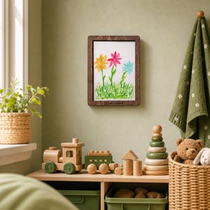 Pode incluir: Uma moldura de madeira retangular contendo um desenho infantil de três flores em amarelo, rosa e azul-claro com caules e folhas verdes, sobre um fundo branco com grama verde na parte inferior, pendurada em uma parede.