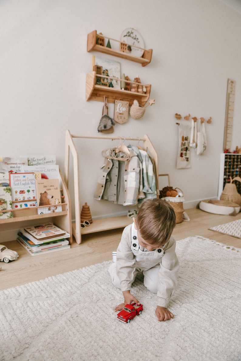 May include: A child plays with a red toy car on a white fluffy rug in a room with a wooden coat rack and shelves. The shelves are decorated with wooden toys and a sign that says "Leo".