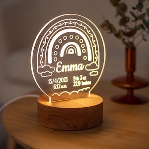 May include: A personalised night light featuring a rainbow design, the name "Emma", and birth details. The light sits on a wooden base. The text includes the date 15/4/2023, 4.19 kg, 58.17 cm, and 6:12 a.m.