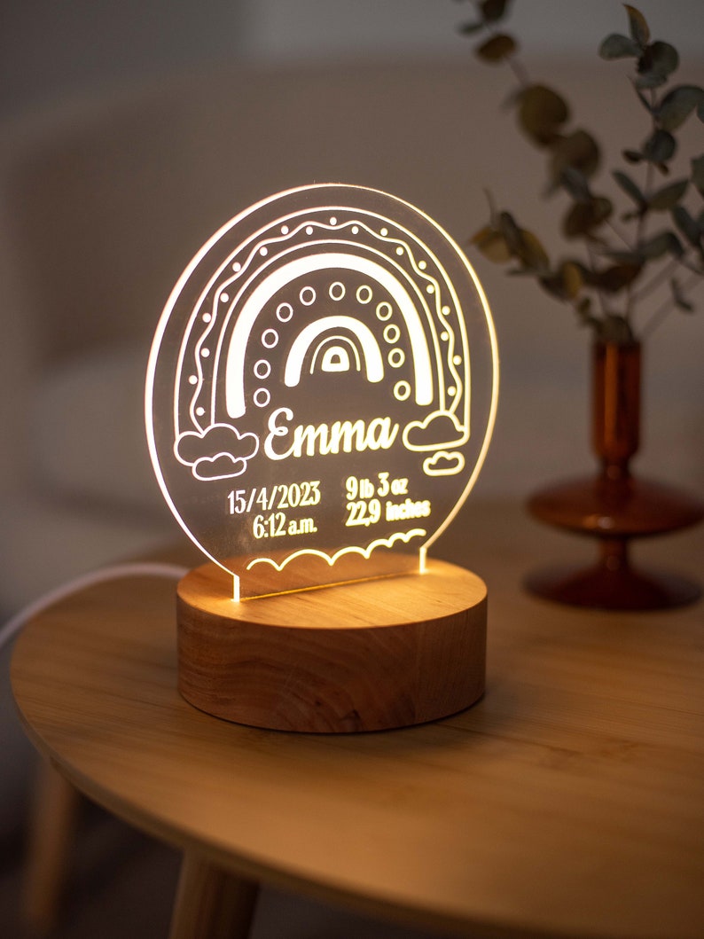May include: A personalised night light featuring a rainbow design, the name "Emma", and birth details. The light sits on a wooden base. The text includes the date 15/4/2023, 4.19 kg, 58.17 cm, and 6:12 a.m.