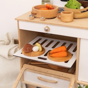 Può includere: Set da cucina giocattolo in legno con finitura bianca e legno naturale. Include un piano cottura, un forno e vari alimenti in legno come carote, funghi e una ciotola di oggetti arancioni. Sono visibili anche una tazza e un cestino di legno.