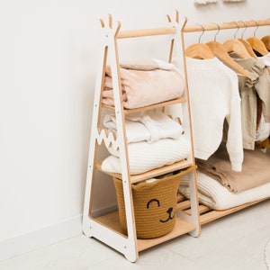 May include: White wooden clothes rack with three shelves and a hanging bar. The rack is shaped like a teepee and has a brown woven basket with a smiling face on the bottom shelf. The rack is filled with folded clothes and blankets.