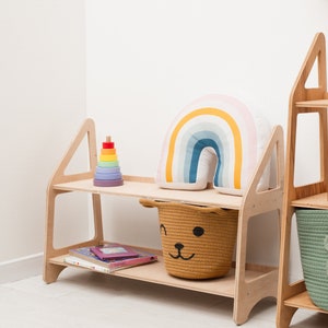 May include: A wooden shelf unit with two shelves. The top shelf has a rainbow-colored pillow and a wooden stacking toy. The bottom shelf has a woven basket with a bear face and a stack of books.