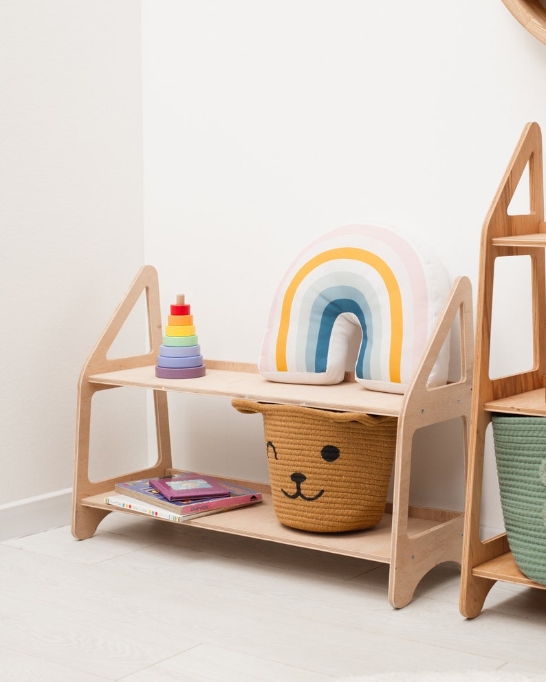 May include: A wooden shelf unit with two shelves. The top shelf has a rainbow-colored pillow and a wooden stacking toy. The bottom shelf has a woven basket with a bear face and a stack of books.