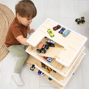 K&ouml;nnte beinhalten: Ein mehrst&ouml;ckiges Spielzeug-Autogarage aus Holz mit Rampe, gef&uuml;llt mit Spielzeugautos. Ein Kind spielt mit einem roten Monstertruck auf der obersten Ebene. Die Garage hat einen wei&szlig;en Rahmen. Die Spielzeugautos sind in verschiedenen Farben.