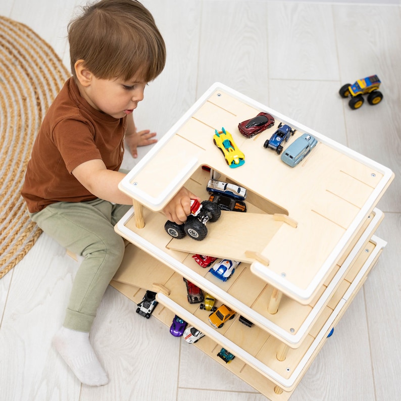K&ouml;nnte beinhalten: Ein mehrst&ouml;ckiges Spielzeug-Autogarage aus Holz mit Rampe, gef&uuml;llt mit Spielzeugautos. Ein Kind spielt mit einem roten Monstertruck auf der obersten Ebene. Die Garage hat einen wei&szlig;en Rahmen. Die Spielzeugautos sind in verschiedenen Farben.