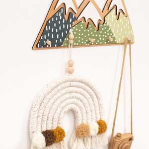 May include: A wooden mountain-shaped wall hook with three hooks and a white and green painted design. A white and brown macrame rainbow with pom-poms hangs from one of the hooks. A wooden camera hangs from another hook.